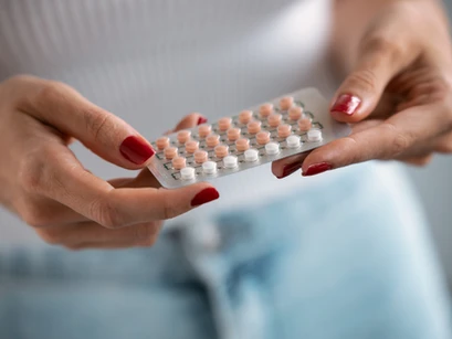 A person holding a blister pack of contraceptive pills with a focus on red nails and casual attire.