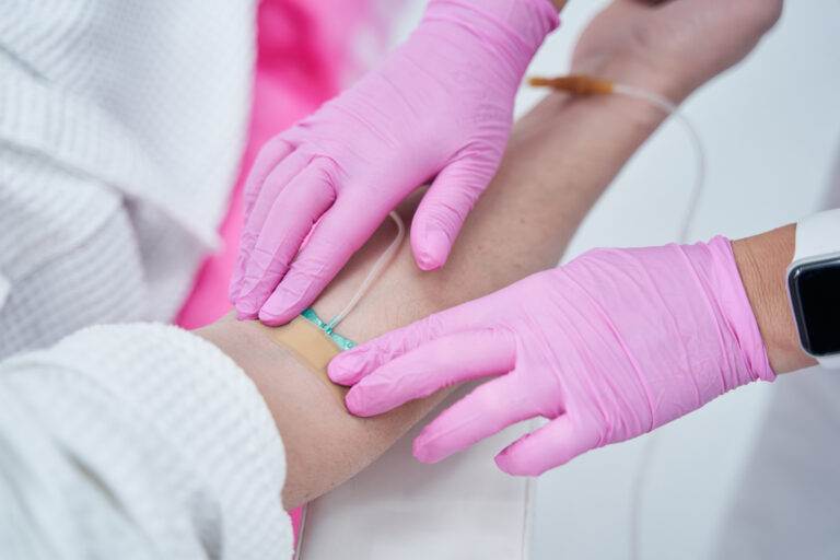 Close-up of a healthcare professional administering an IV infusion to a patient's arm, wearing pink gloves.