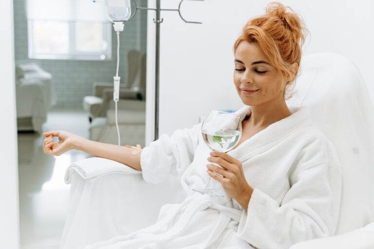 Woman receiving IV therapy while sitting comfortably and drinking a glass of water.