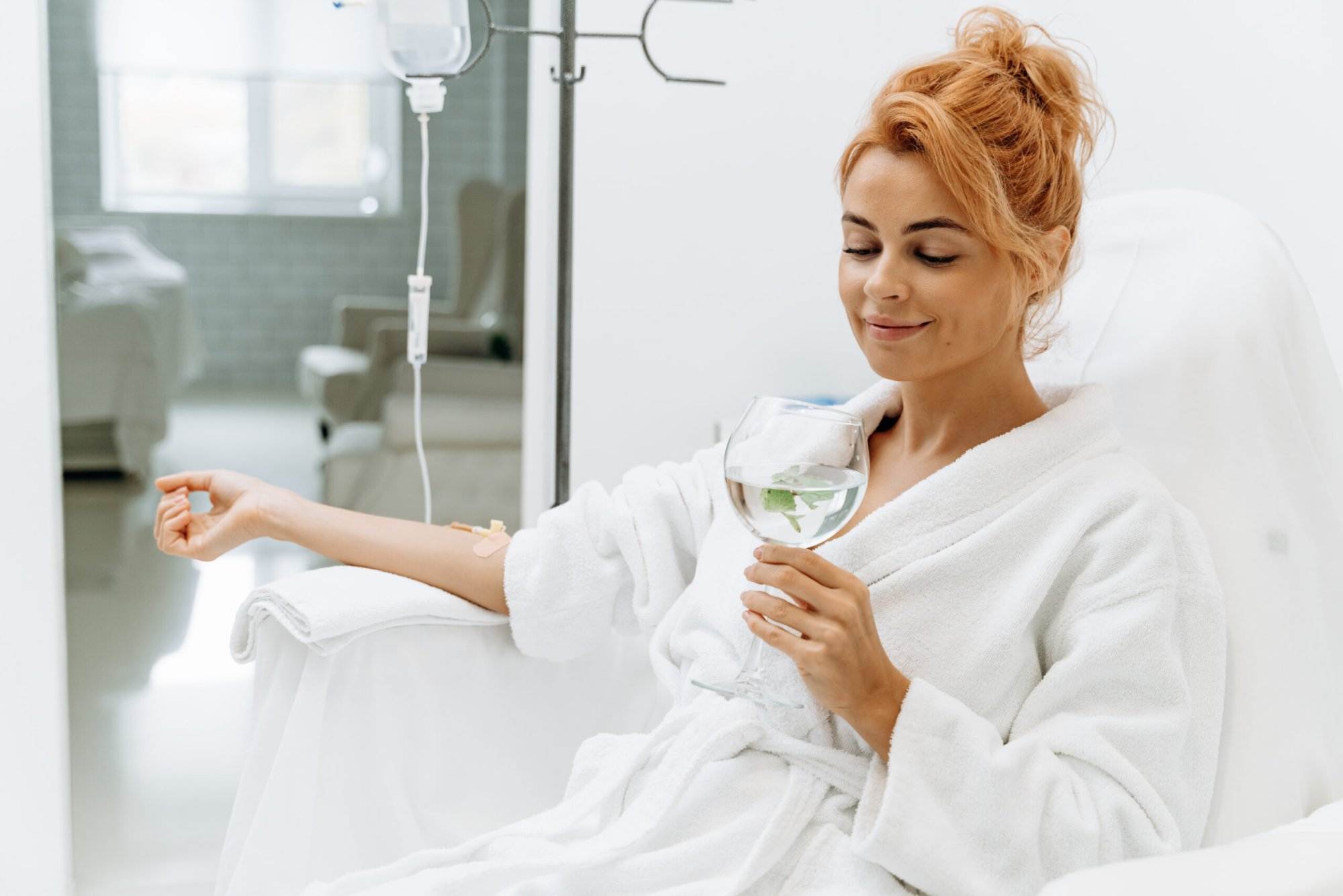 Woman receiving IV therapy while sitting comfortably and drinking a glass of water.