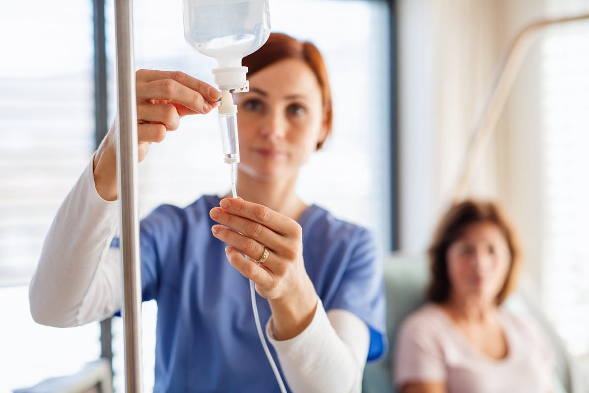 A nurse in blue scrubs carefully adjusts an IV infusion, with a patient seated in the background. The scene is set in a brightly lit medical facility.