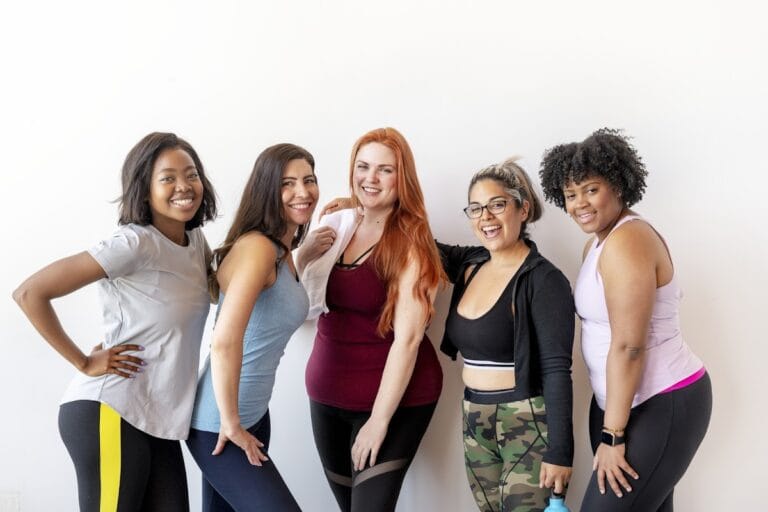 Diverse group of women smiling at the gym in workout attire, showcasing team spirit and fitness.