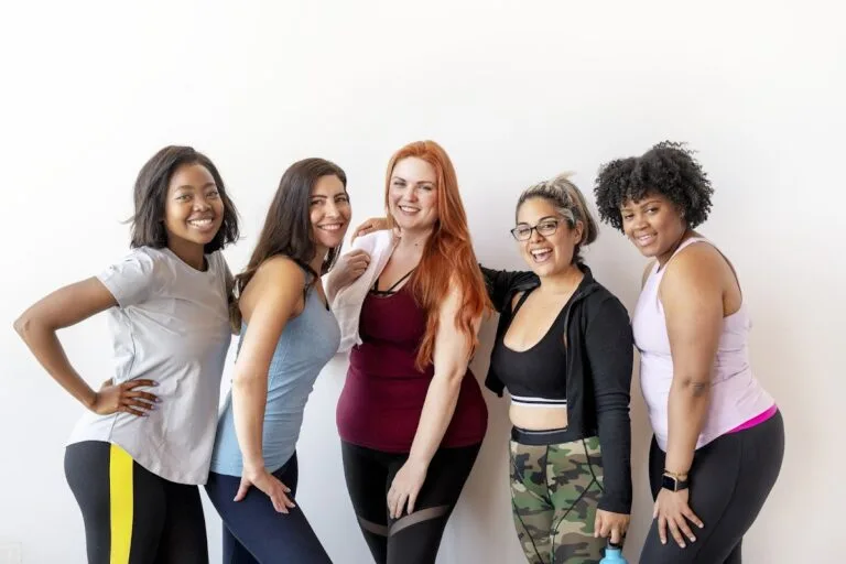 Diverse group of women smiling at the gym in workout attire, showcasing team spirit and fitness.